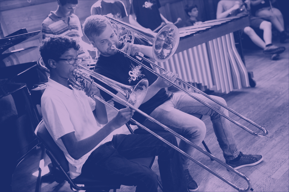 A purple filter, of a male student and male teacher, sitting down next to each other, performing on the trombone.